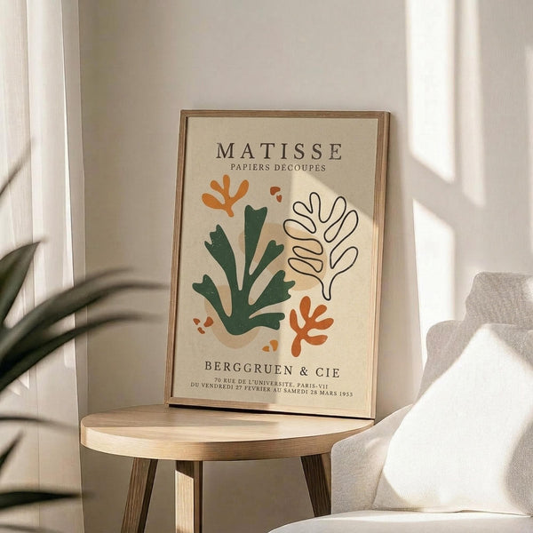 May include: Framed art print in thin wooden frame displaying abstract cut-out style botanical shapes in green, orange, and cream colors with French text, positioned on round wooden side table next to beige upholstered chair and plant leaves, with natural sunlight casting shadows on neutral wall.