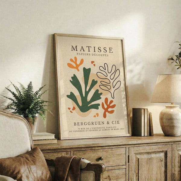 May include: Framed Matisse-inspired poster with abstract botanical cutout shapes in green, orange and beige on cream background, displayed on rustic wooden sideboard alongside fern plant, books, ceramic vase and brown leather chair in neutral-toned living room setting