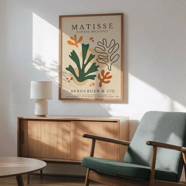 May include: Framed poster featuring Matisse-style cut-out botanical shapes in green and orange on cream background, displayed above a mid-century modern wooden sideboard with white ceramic lamp. Green upholstered armchair with wooden frame sits nearby in sunlit room with white walls.