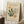 May include: A cream-colored vintage exhibition poster featuring abstract botanical cutout shapes in green, orange, and white displayed in a light wooden frame. The poster leans against white paneled walls on a textured white rug, with a coffee cup on a wooden block and beige fabric visible nearby.
