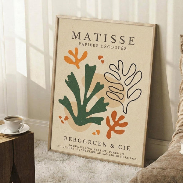 May include: A cream-colored vintage exhibition poster featuring abstract botanical cutout shapes in green, orange, and white displayed in a light wooden frame. The poster leans against white paneled walls on a textured white rug, with a coffee cup on a wooden block and beige fabric visible nearby.