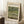 May include: Framed exhibition poster featuring Monet's Japanese Footbridge painting displayed in a bright living room corner. The poster shows a curved blue-green bridge over a lily pond with lush vegetation. Light wood frame leans against white paneled walls next to a beige armchair and wooden side table with white coffee cup. Cream-colored poster has museum-style typography with artwork title and 1899 date.