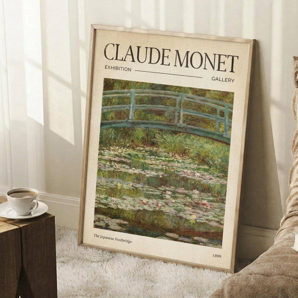 May include: Framed exhibition poster featuring Monet's Japanese Footbridge painting displayed in a bright living room corner. The poster shows a curved blue-green bridge over a lily pond with lush vegetation. Light wood frame leans against white paneled walls next to a beige armchair and wooden side table with white coffee cup. Cream-colored poster has museum-style typography with artwork title and 1899 date.