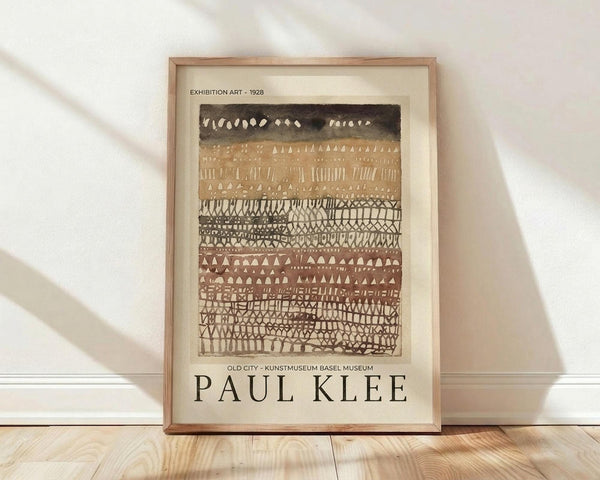 May include: Framed art print leaning against white wall on light wood floor. Abstract artwork features horizontal bands of geometric patterns in earth tones - black at top, warm browns and tans below. Intricate tribal-style motifs including triangles, lines, dots, and maze-like designs fill each striped section. Natural wood frame surrounds cream-colored print with artist name at bottom. Soft natural lighting creates subtle shadows on textured white wall.