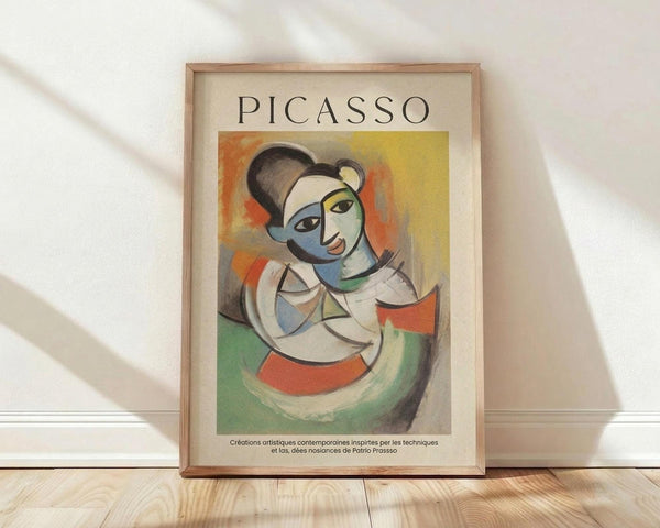 May include: Framed poster print in light wood frame featuring cubist-style portrait of woman with geometric facial features in blue, green, yellow and orange tones. French text below artwork reads about contemporary artistic creations inspired by Picasso's techniques. Frame leans against white wall on light wooden floor with soft natural lighting creating diagonal shadows.
