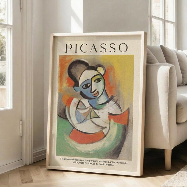 May include: Framed Picasso-style cubist portrait poster showing abstract figure with geometric face in blues, oranges, yellows and greens, leaning against beige armchair in bright living room with white walls and large windows