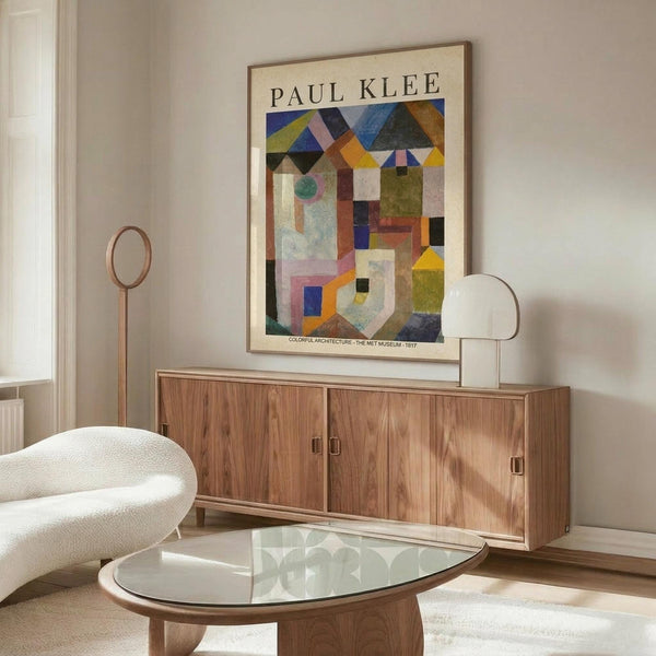 May include: Paul Klee abstract geometric artwork poster displayed above wooden credenza in modern living room. Colorful architectural composition features triangular rooftops, rectangular buildings, and circular elements in blues, yellows, oranges, and greens. White curved chair and glass oval coffee table visible in foreground. Poster appears framed in thin wood frame against light gray wall.