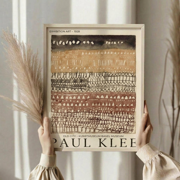 May include: Person in cream sweater holding framed poster against white curtained window, with dried wheat stems visible. The artwork features horizontal bands of geometric patterns in earth tones - black, tan, rust, and cream - with triangles, dots, lines and crosshatch designs. Light wooden frame surrounds the abstract composition.