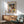 May include: Paul Klee abstract geometric art print in wooden frame hanging above modern wood sideboard. Colorful cubist composition features triangular houses, architectural forms in blues, yellows, oranges, and greens. Mid-century modern room setting with mint green upholstered armchair, white ceramic table lamp, and natural sunlight creating shadows on white wall.
