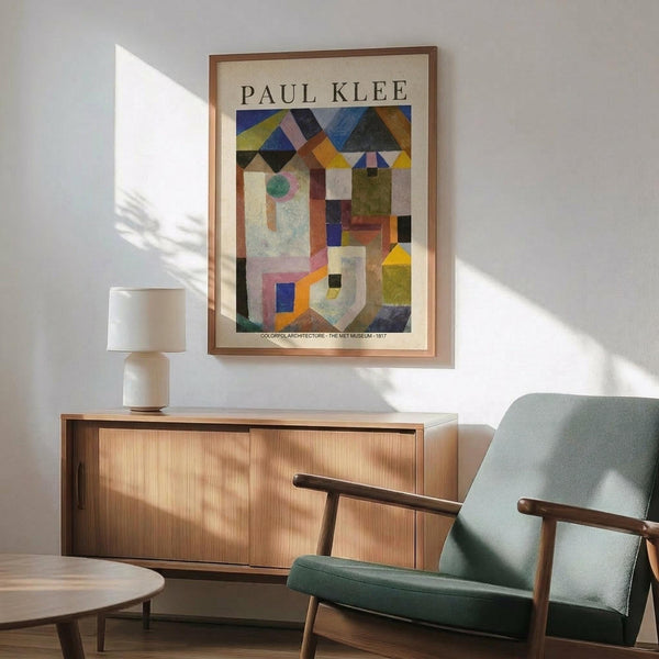 May include: Paul Klee abstract geometric art print in wooden frame hanging above modern wood sideboard. Colorful cubist composition features triangular houses, architectural forms in blues, yellows, oranges, and greens. Mid-century modern room setting with mint green upholstered armchair, white ceramic table lamp, and natural sunlight creating shadows on white wall.