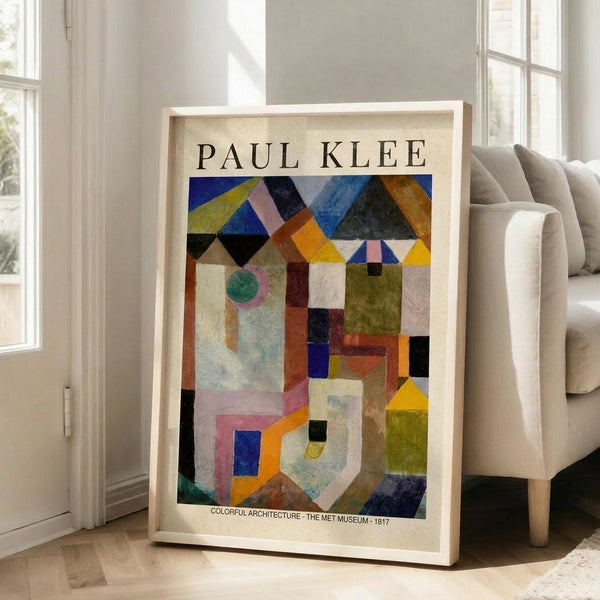May include: Framed Paul Klee abstract art print featuring geometric shapes in vibrant colors including blues, yellows, oranges, and greens arranged in an architectural composition. The print shows museum exhibition text at bottom and is displayed in a light wood frame leaning against a white wall next to a beige upholstered chair in a bright, modern living room with large windows.