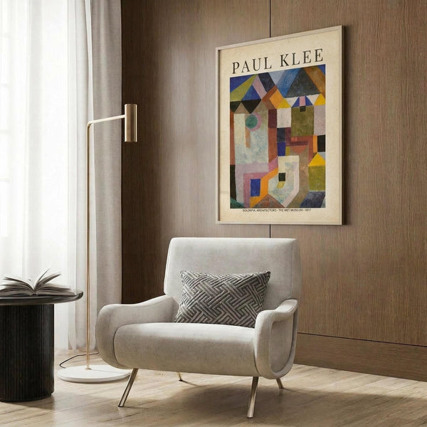 May include: Paul Klee abstract art poster featuring colorful geometric buildings and houses in blues, oranges, yellows and earth tones, displayed in cream matted frame on wood paneled wall above modern beige upholstered armchair with geometric patterned pillow, beside brass floor lamp and dark round side table with open book in contemporary living room setting