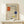 May include: Framed art print featuring geometric grid composition with red, blue, and yellow rectangular blocks on cream background, displayed on light wood surface beside small white wrapped gift with natural twine and pine sprig against neutral wall