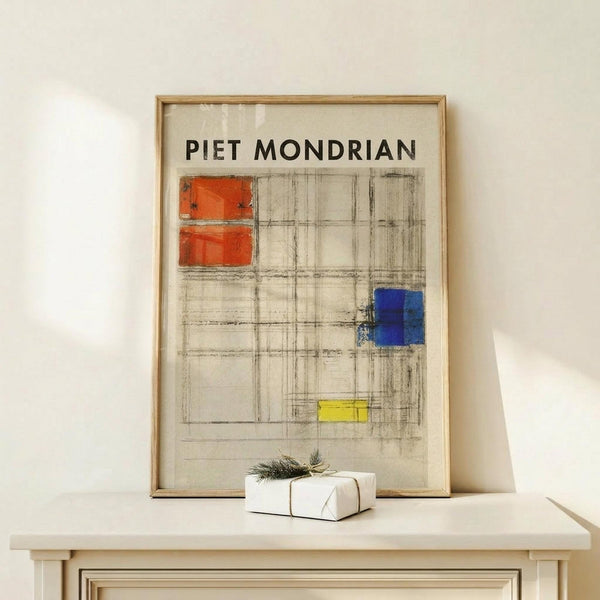 May include: Framed art print featuring geometric grid composition with red, blue, and yellow rectangular blocks on cream background, displayed on light wood surface beside small white wrapped gift with natural twine and pine sprig against neutral wall