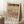 May include: Framed Paul Klee exhibition poster leaning against white wall on textured cream rug. Abstract artwork features horizontal bands of geometric patterns in earth tones - dark green at top, warm ochre middle, terracotta and brown lower sections with triangular and linear motifs. Light wood frame, cream matting with black text. Small wooden side table with white coffee cup visible on left. Cozy home interior setting with soft natural lighting.