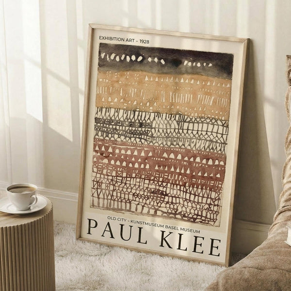 May include: Framed Paul Klee exhibition poster leaning against white wall on textured cream rug. Abstract artwork features horizontal bands of geometric patterns in earth tones - dark green at top, warm ochre middle, terracotta and brown lower sections with triangular and linear motifs. Light wood frame, cream matting with black text. Small wooden side table with white coffee cup visible on left. Cozy home interior setting with soft natural lighting.