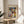 May include: Paul Klee abstract art poster in thin wooden frame displayed on round wooden side table next to beige upholstered chair. The colorful geometric artwork features overlapping shapes in blues, yellows, greens, and earth tones arranged like architectural forms. Soft natural lighting creates shadows across white walls, with green plant leaves visible in corner of minimalist room setting.