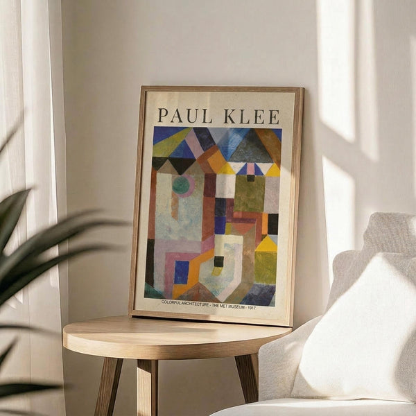 May include: Paul Klee abstract art poster in thin wooden frame displayed on round wooden side table next to beige upholstered chair. The colorful geometric artwork features overlapping shapes in blues, yellows, greens, and earth tones arranged like architectural forms. Soft natural lighting creates shadows across white walls, with green plant leaves visible in corner of minimalist room setting.