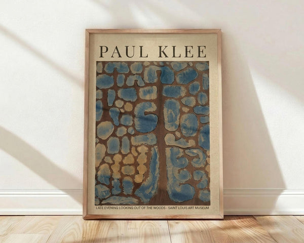 May include: Framed art print featuring Paul Klee's abstract artwork with organic blue and beige shapes on brown background, displayed in light wood frame against white wall on wooden floor with soft natural lighting creating diagonal shadows.