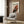 May include: Framed Pollock abstract art poster with red, black and white paint splatters hanging on wood panel wall above beige upholstered chair with geometric patterned pillow, brass floor lamp, and dark round side table with open book in modern living room setting