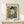 May include: Framed Picasso-style cubist portrait poster showing a figure in dark hat and blue clothing with white collar against colorful geometric background, displayed on light wood console table beside small wrapped gift, in minimalist room setting with cream walls and natural lighting.