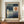 May include: Framed Paul Klee exhibition poster displayed on wooden sideboard featuring abstract geometric composition with blue-green rectangular bands, orange circle resembling sun, and pale crescent moon shape. Poster shows 1922 Yale University Art Gallery exhibition text. Styled with small white ceramic vase and stacked books against cream wall with white trim molding.