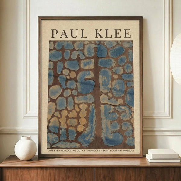 May include: Framed Paul Klee art print displayed on wooden credenza against cream walls with white trim. The abstract artwork features organic blue and beige shapes resembling stones or cellular forms on brown background. Simple white ceramic vase and stacked books sit alongside the medium-sized framed print in warm, minimalist interior setting.