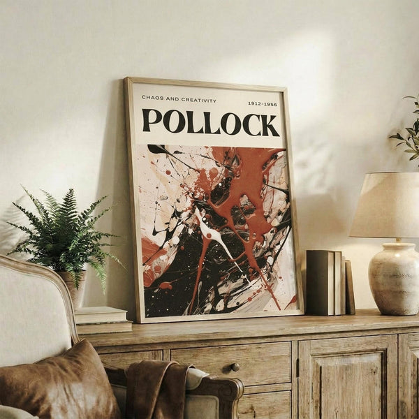 May include: Framed art print featuring abstract expressionist painting with bold black, red, and white paint splatters and drips displayed on rustic wooden sideboard in modern living room setting alongside fern plant, ceramic vase, and stacked books with warm natural lighting and neutral beige walls.