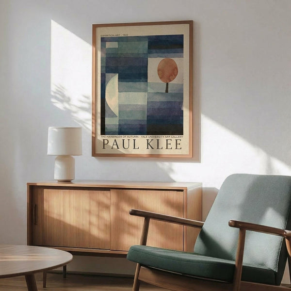 May include: Paul Klee abstract art poster in wooden frame hanging above mid-century modern wooden sideboard with table lamp. Mint green upholstered armchair with wooden arms sits nearby. The artwork features geometric shapes in blues, greens, and orange tones depicting stylized tree forms. Warm sunlight streams across white walls creating dramatic shadows in this contemporary living room setting.