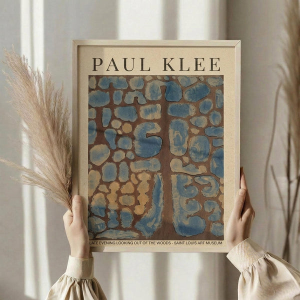 May include: Person in beige ruffled sleeves holding framed Paul Klee art print featuring abstract blue and brown stone-like shapes on warm background. Natural pampas grass stems visible beside frame. Soft window lighting creates neutral, minimalist styling against light curtains.