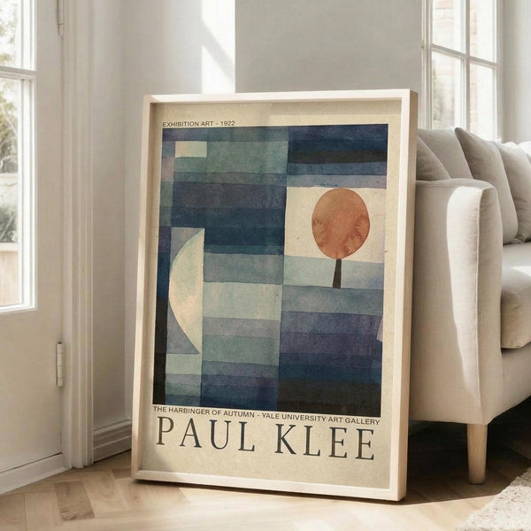 May include: Framed Paul Klee exhibition poster leaning against beige sofa in bright living room with white walls and windows. Abstract artwork features geometric blue and teal rectangular blocks with white crescent moon and orange-red circle, resembling a landscape. Light wood frame on hardwood floor with natural sunlight streaming in.
