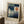 May include: A framed Paul Klee exhibition poster leaning against a white wall on a textured white rug. The artwork features geometric blue and teal rectangular shapes with a white crescent moon on the left and orange-red circle on the right, resembling an abstract autumn landscape. A wooden side table with white coffee cup and saucer sits nearby, with soft natural lighting creating window shadows on the wall.
