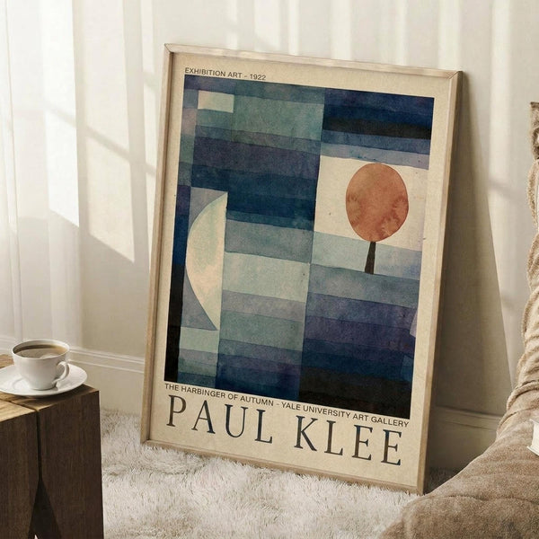May include: A framed Paul Klee exhibition poster leaning against a white wall on a textured white rug. The artwork features geometric blue and teal rectangular shapes with a white crescent moon on the left and orange-red circle on the right, resembling an abstract autumn landscape. A wooden side table with white coffee cup and saucer sits nearby, with soft natural lighting creating window shadows on the wall.