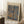 May include: Framed art poster leaning against white wall corner on fluffy cream rug. Shows abstract mosaic-style artwork with irregular blue and beige shapes on brown background, resembling stones or organic forms. Light wood frame, museum exhibition style with artist name at top. Small wooden side table with white coffee cup visible on left. Soft natural lighting creates shadows on wall.