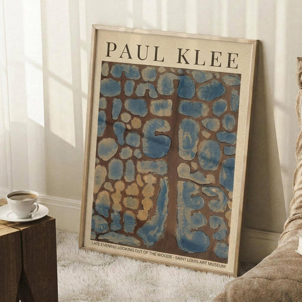 May include: Framed art poster leaning against white wall corner on fluffy cream rug. Shows abstract mosaic-style artwork with irregular blue and beige shapes on brown background, resembling stones or organic forms. Light wood frame, museum exhibition style with artist name at top. Small wooden side table with white coffee cup visible on left. Soft natural lighting creates shadows on wall.