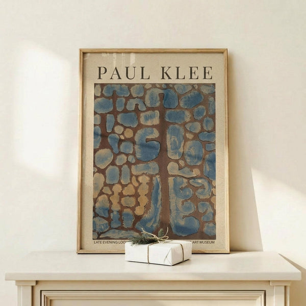 May include: Framed Paul Klee art poster featuring abstract organic shapes in blue and brown tones, displayed on light wood console table beside small white wrapped gift with twine and greenery sprig against cream wall