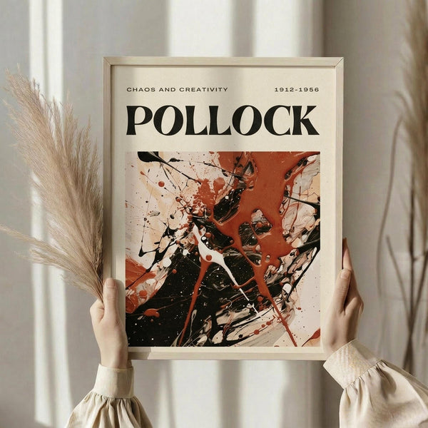 May include: Person in beige ruffled sleeves holding framed abstract art poster with black paint splatters and red drips on cream background, featuring "POLLOCK" text and dates "1912-1956." Light wood frame against white curtained window with dried pampas grass visible to left side.