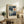 May include: Framed Paul Klee exhibition poster featuring abstract geometric composition with blue and teal rectangular blocks, white crescent moon, and orange circular tree form on cream background. Displayed on rustic wooden sideboard alongside fern plant, stacked books, ceramic vessels, and beige upholstered chair in warm, naturally-lit living room setting.