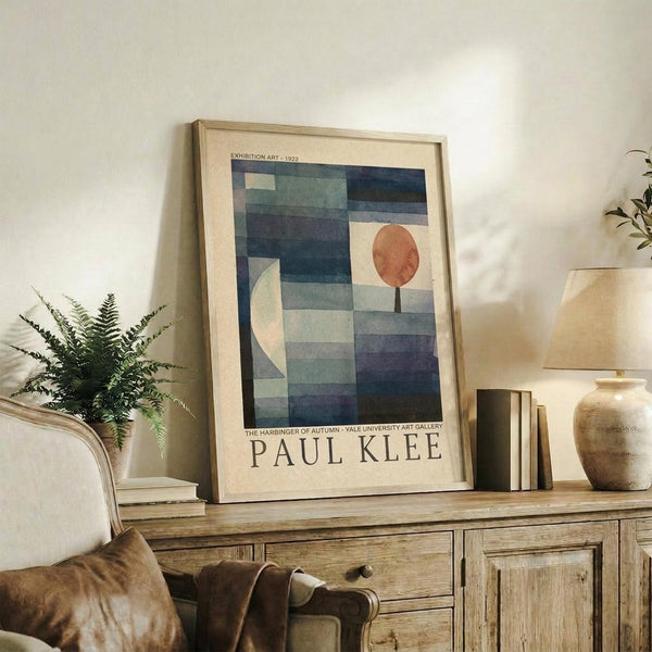 May include: Framed Paul Klee exhibition poster featuring abstract geometric composition with blue and teal rectangular blocks, white crescent moon, and orange circular tree form on cream background. Displayed on rustic wooden sideboard alongside fern plant, stacked books, ceramic vessels, and beige upholstered chair in warm, naturally-lit living room setting.