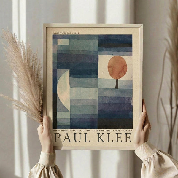 May include: Hands holding a framed vintage exhibition poster featuring Paul Klee's geometric abstract artwork with blue rectangular blocks, white crescent moon, and orange circle with black stem. Light wood frame against neutral curtain backdrop with dried wheat stems visible on left side.