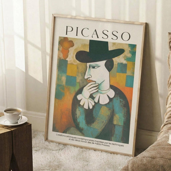 May include: Framed Picasso-style portrait print leaning against white wall in cozy room setting. Artwork shows figure in dark hat and blue clothing with white collar against colorful geometric background. Light wood frame, positioned on textured cream rug next to wooden side table with coffee cup and beige chair arm visible.