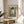 May include: Framed Picasso-style portrait print featuring a figure in a dark hat and blue-green clothing with white collar against a colorful geometric background. The artwork sits on a wooden round side table next to a beige upholstered chair, with natural sunlight streaming across white walls and a plant visible in the corner of the modern living space.