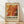 May include: Abstract exhibition poster in wooden frame leaning against white wall on light wood floor. Features flowing marbled paint in warm oranges, reds, and yellows with black accents and white dots. Vintage-style typography reads "POLLOCK" and "EMBRACE THE CHAOTIC REALM OF CREATIVITY" with exhibition dates. Soft natural lighting creates diagonal shadows across the scene.