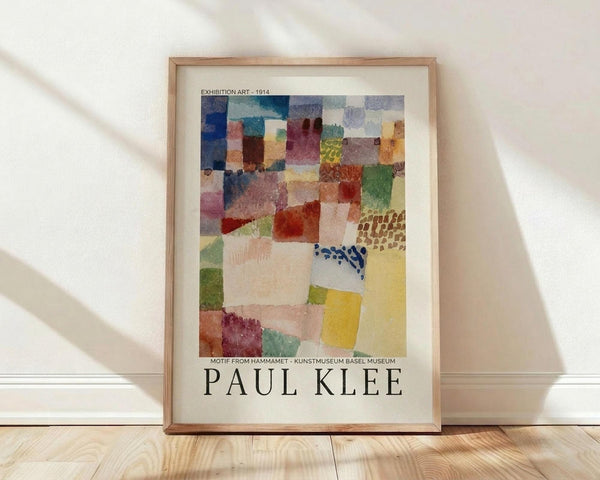 May include: Framed exhibition poster featuring Paul Klee's abstract watercolor artwork from 1914, displayed against a white wall on wooden flooring. The artwork shows geometric patches in blues, reds, yellows, greens, and earth tones with various textures and patterns. Natural light creates soft shadows across the scene. The poster has museum exhibition text and the artist's name in bold letters below the colorful abstract composition.