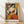 May include: Framed cubist-style art print featuring a woman in geometric forms holding a colorful fan, rendered in orange, yellow, white, and dark tones. The wooden-framed poster leans against a white wall on light wood flooring with natural shadows, showing "PICASSO" text at bottom.