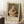 May include: Framed exhibition poster featuring Renoir's 1875 painting of a woman in white dress holding a tabby cat, with cream matting and light wood frame. Poster leans against white wall on textured white rug next to wooden side table with coffee cup and saucer. Soft natural lighting creates gentle shadows across the scene.