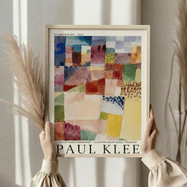 May include: Person in cream blouse holding framed Paul Klee exhibition poster featuring abstract watercolor composition of overlapping geometric shapes in blues, reds, yellows, greens, and purples. Light wooden frame against white curtained window with dried wheat stalks visible on left side.