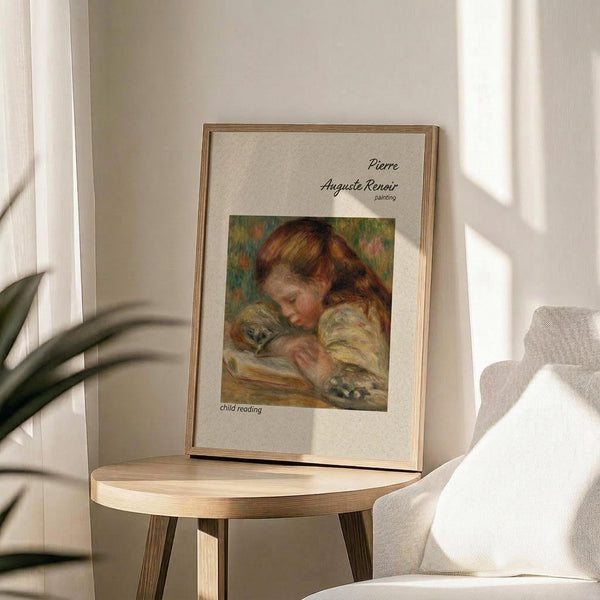 May include: Framed art print displaying Pierre Auguste Renoir's impressionist painting of a young child with reddish-brown hair reading a book. The reproduction shows soft, warm brushstrokes in peachy and golden tones against a muted background. Print sits on a round wooden side table next to a beige upholstered chair, with natural sunlight casting shadows and a plant visible in the corner of the room.