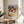 May include: Framed art print featuring Paul Klee's colorful abstract watercolor composition with geometric squares and rectangles in blues, reds, yellows, and greens, displayed on a round wooden side table next to a beige upholstered chair. Natural sunlight streams across the neutral-toned room setting with a plant visible in the corner.
