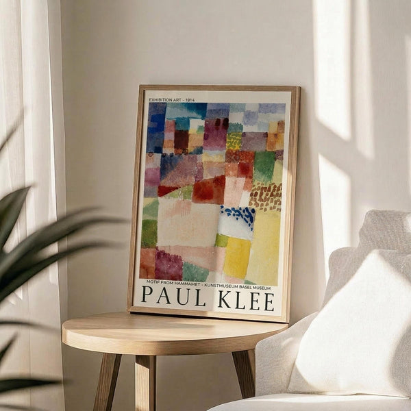 May include: Framed art print featuring Paul Klee's colorful abstract watercolor composition with geometric squares and rectangles in blues, reds, yellows, and greens, displayed on a round wooden side table next to a beige upholstered chair. Natural sunlight streams across the neutral-toned room setting with a plant visible in the corner.